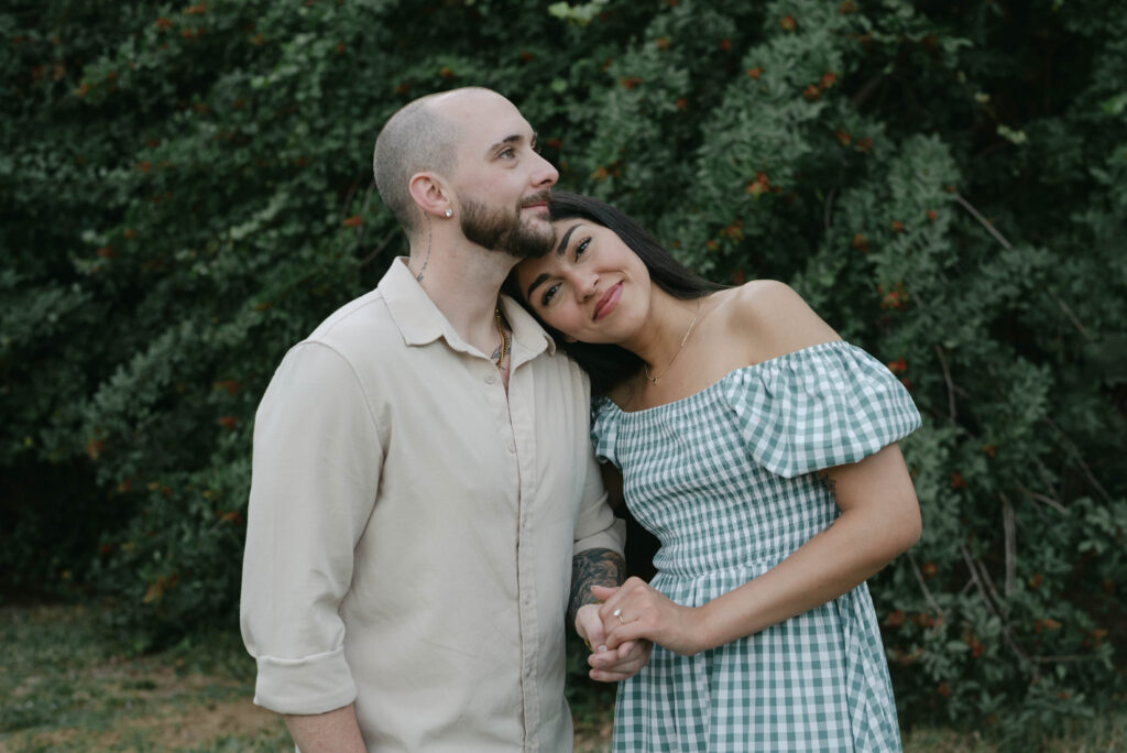 Bride leans on the groom's shoulder and smiles at the camera during backyard proposal